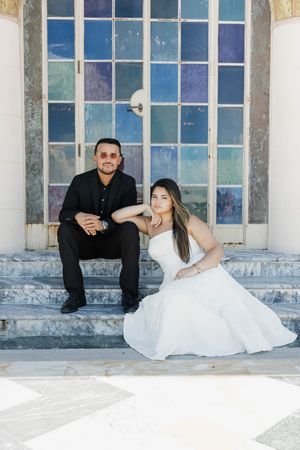 Bride in white gown and groom in black suit sitting on marble steps before a colorful stained glass window.