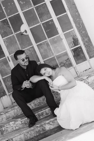 Black and white photo of a bride and groom sitting on steps, she rests her head on his hands.