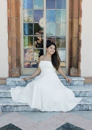 A bride in a strapless white ball gown sits gracefully on marble steps in front of an ornate window.