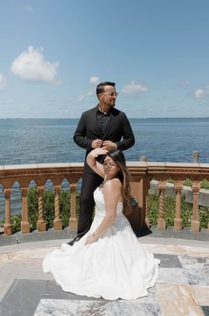 Bride in white gown sits as groom in black tuxedo stands behind her on elegant terrace overlooking ocean.
