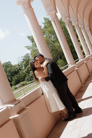 Couple in wedding attire embracing between elegant pink columns of a classical colonnade with lush green trees in background.