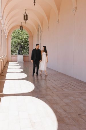 Couple walking through elegant pink arched colonnade with sunlight casting arch shadows on tiled floor.