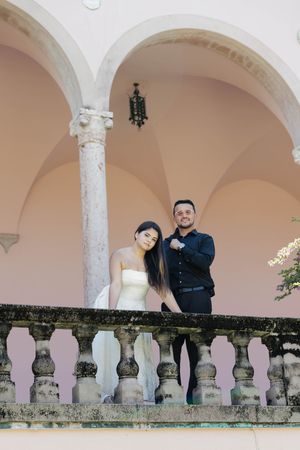 Bride and groom posing on a balcony with arched architecture and ornate stone railings.