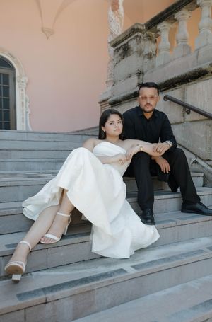 Bride in white strapless gown and groom in black sit together on stone steps of elegant building.