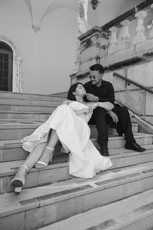 Black and white photo of a couple sitting on marble steps, bride in white dress leaning on groom in black suit.
