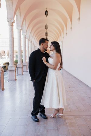 Couple in elegant attire sharing a romantic moment under arched colonnade, woman in white dress, man in black suit.