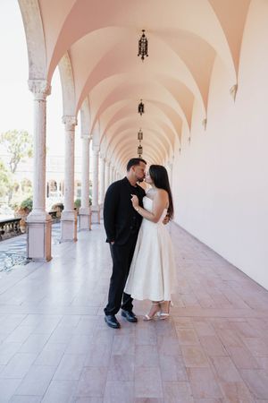 Couple in wedding attire sharing a romantic moment in an elegant arched colonnade corridor.