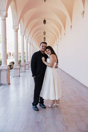 Couple in wedding attire embracing in elegant arched colonnade, groom in black suit, bride in white midi dress.