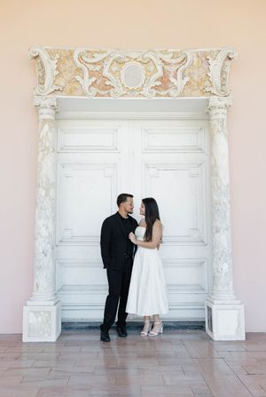Couple in elegant attire standing before ornate white baroque door with decorative columns and intricate molding.