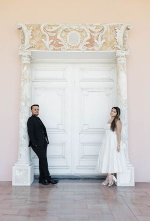 Couple in wedding attire posing on either side of an ornate white double door with decorative columns.