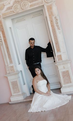 Bride in white gown sits on floor as groom in black stands in ornate white doorway during elegant wedding photo session.