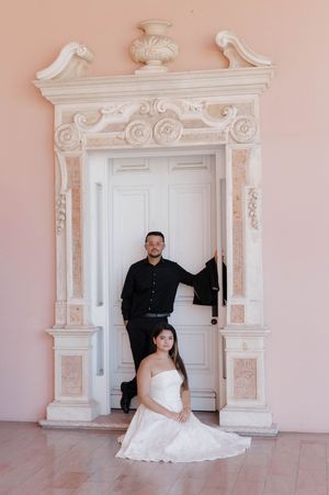 Bride and groom posing at ornate white doorway against pink wall in wedding portraits.