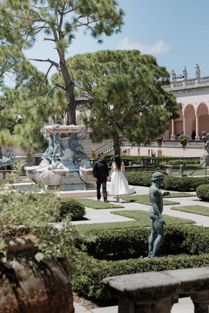 Couple in wedding attire poses near ornate fountain and bronze sculptures in a formal Italian garden.