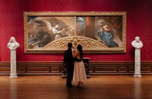 Couple embracing before a grand classical painting in an elegant red-walled art museum gallery with marble busts.