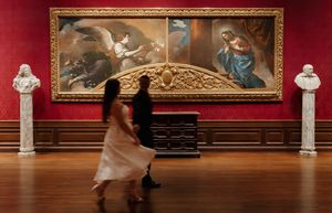 Two visitors walk past a large classical painting in a grand red-walled gallery with marble busts on pedestals.