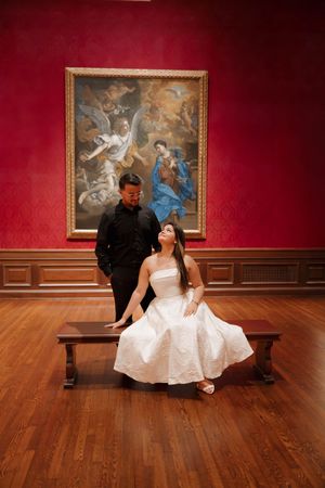Couple poses in art museum gallery with red walls and classical painting, woman in white dress seated on bench.
