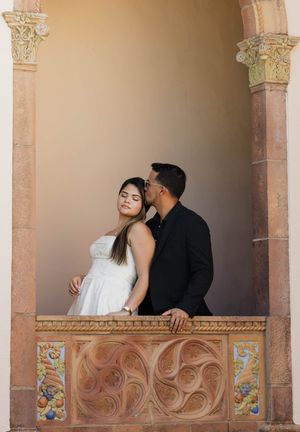 Couple in romantic pose on ornate balcony, woman in white dress, man in black shirt kissing her cheek.