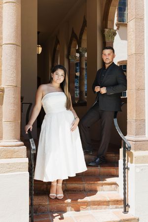 Bride in white midi dress and groom in black suit posing on stone steps between columns.