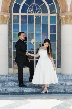 Couple posing on marble steps before ornate stained glass window, man in black suit, woman in white dress.
