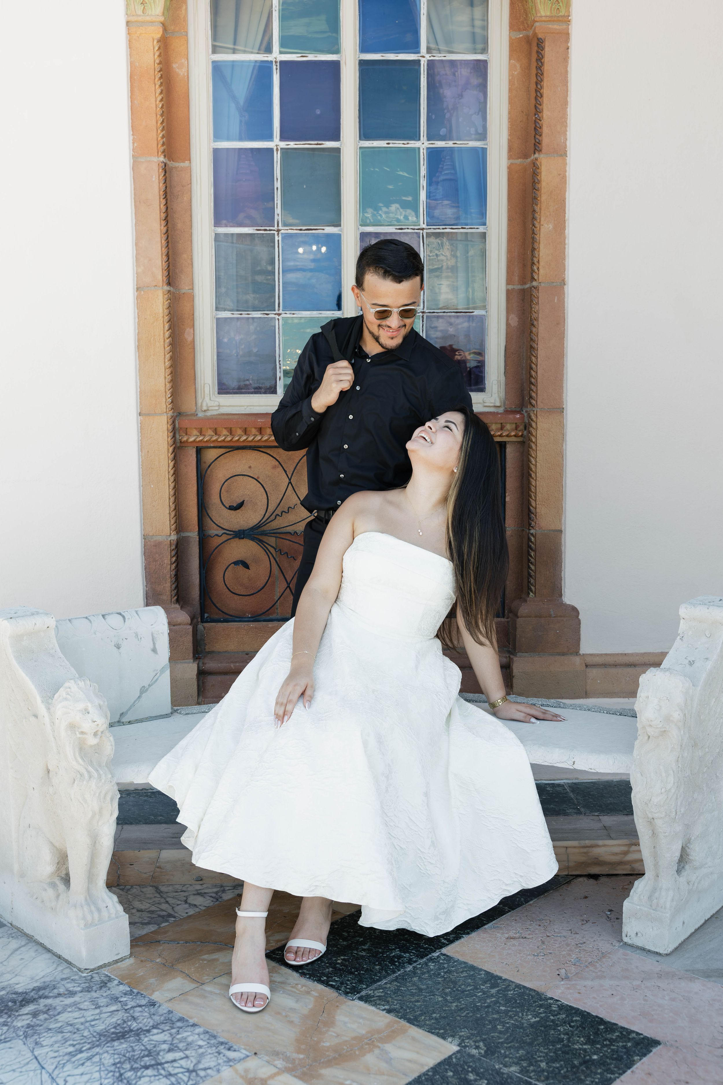 Couple posing elegantly outside a historic building, woman in white dress, man in black shirt with sunglasses.