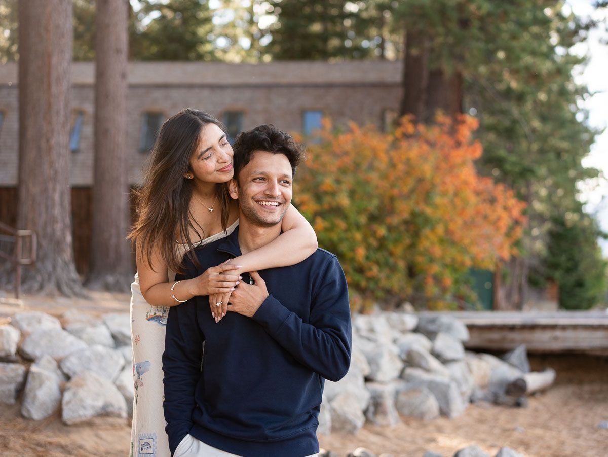 A happy couple embraces outdoors near a rustic stone building surrounded by pine trees and colorful autumn foliage.