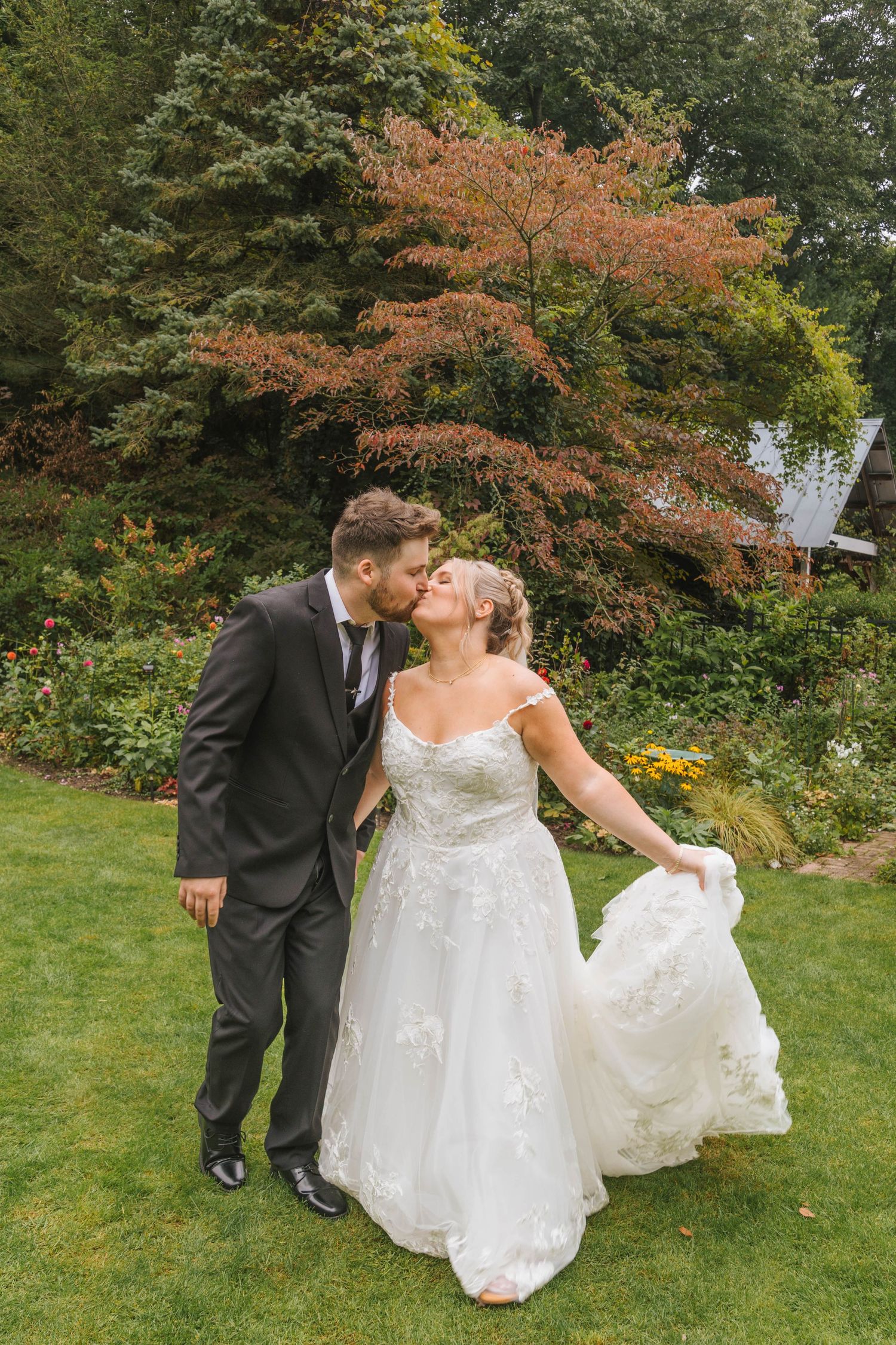 Bride and groom share a kiss outdoors on green lawn with colorful autumn trees in background at wedding.