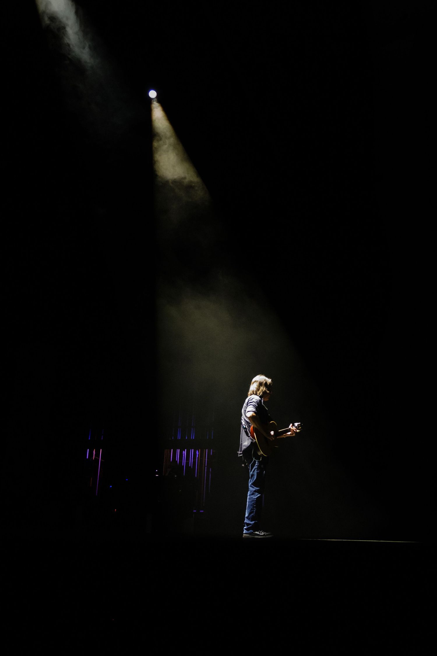 A silhouetted guitar player stands beneath a spot light during a performance.