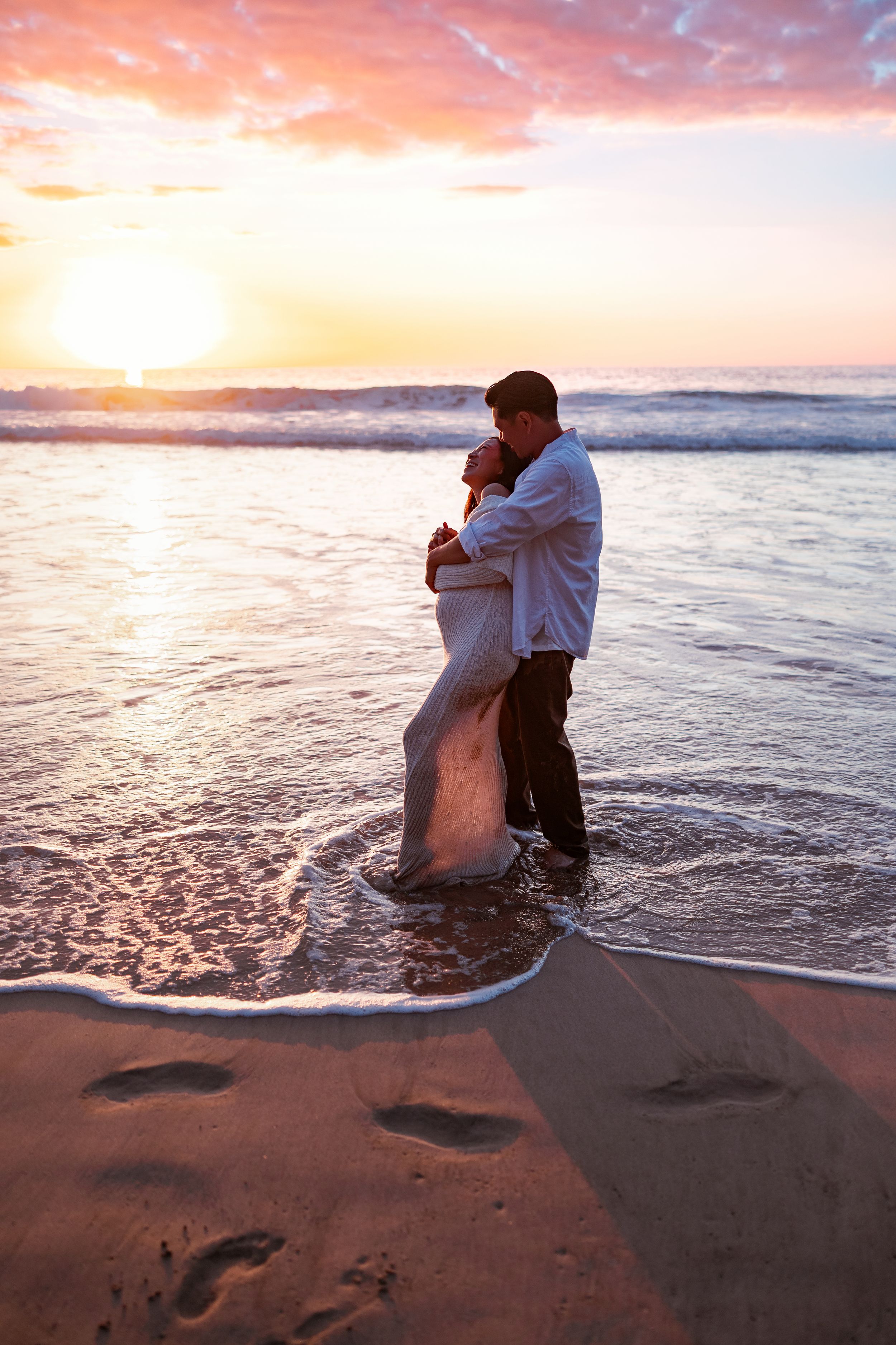 Happy couple embracing and laughing on a sandy beach at sunset, standing in shallow ocean waves.