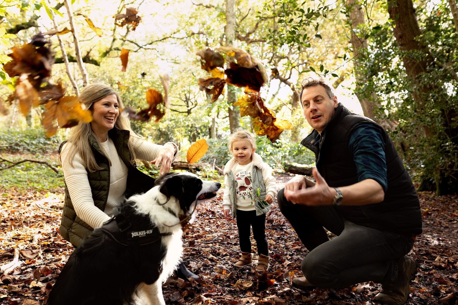Happy family with baby posing outdoors in autumn woodland setting beneath a large oak tree.