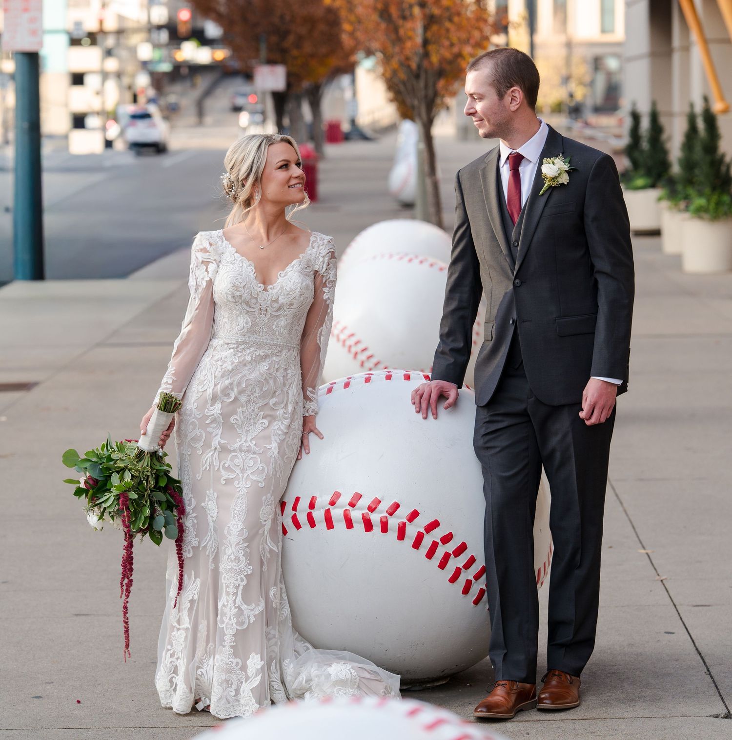 Bride and groom posing beside a giant baseball statue on a city sidewalk, bride holding a bouquet in a lace gown.