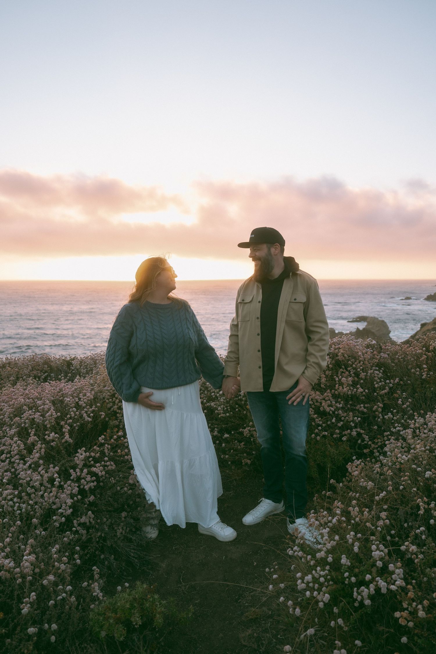 A couple sits on a beach at sunset, wearing matching “mom” and “dad” baseball caps, embracing romantically.