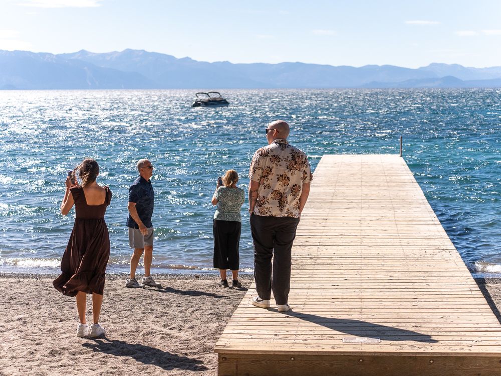 Four people stand on a sunny beach dock overlooking sparkling blue Lake Tahoe with mountains in the background.