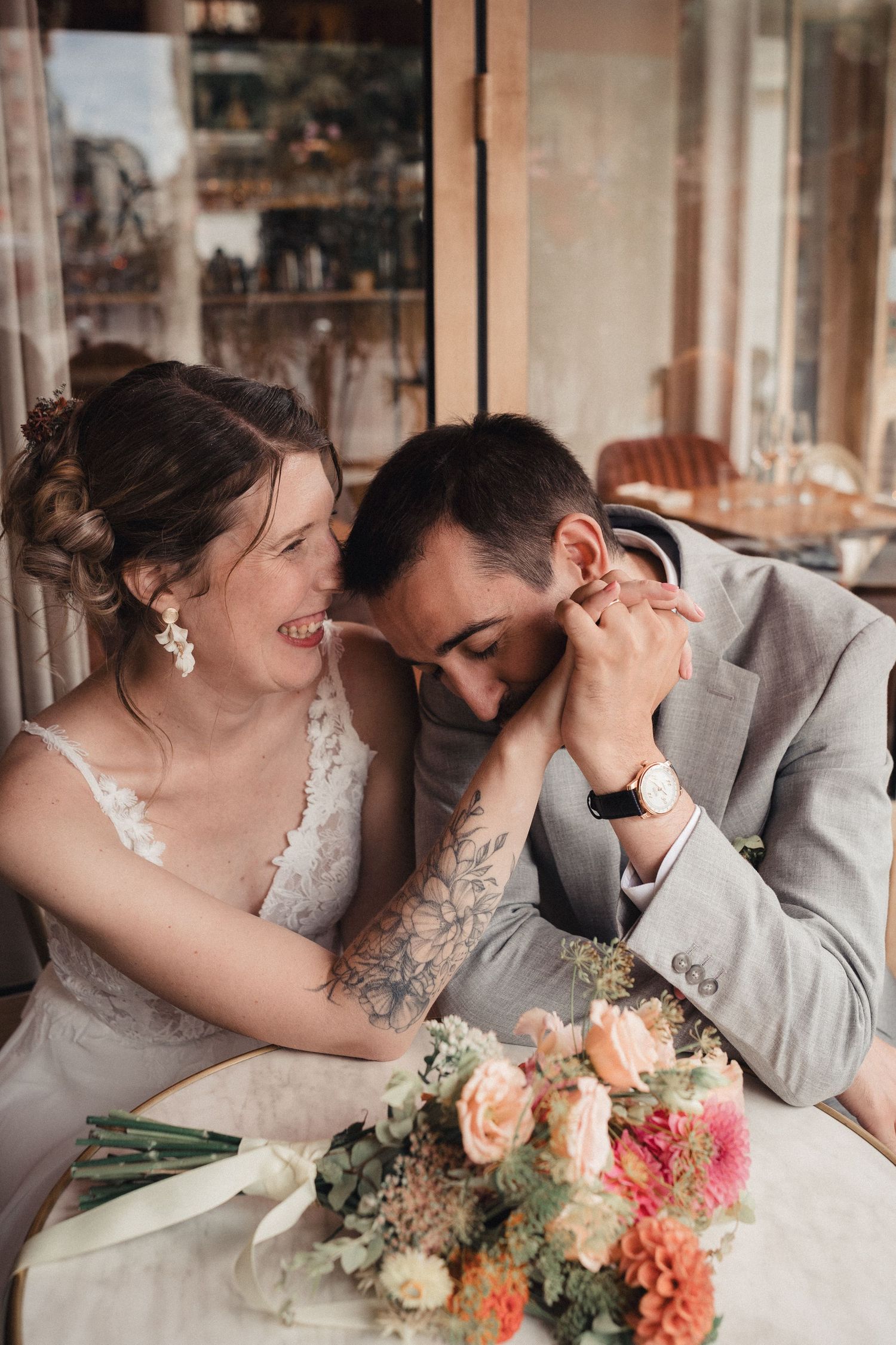 A joyful bride and groom share an intimate moment at a table, with a colorful floral bouquet in the foreground.