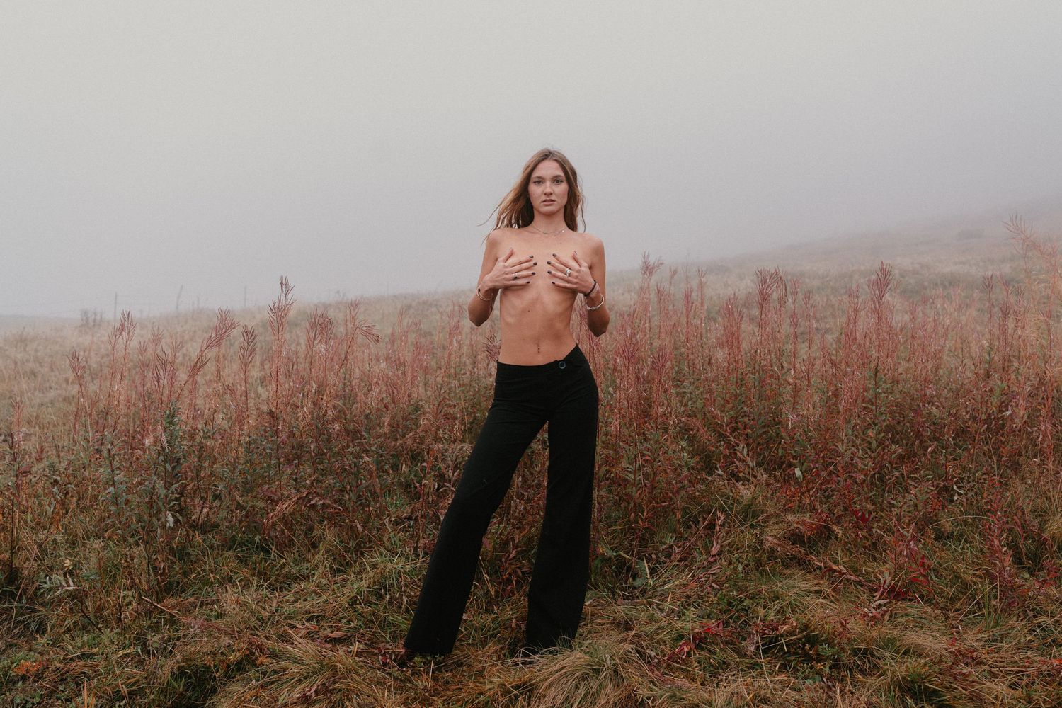 Woman in black wide-leg pants standing topless in a misty field with tall pink wildflowers on a foggy day.