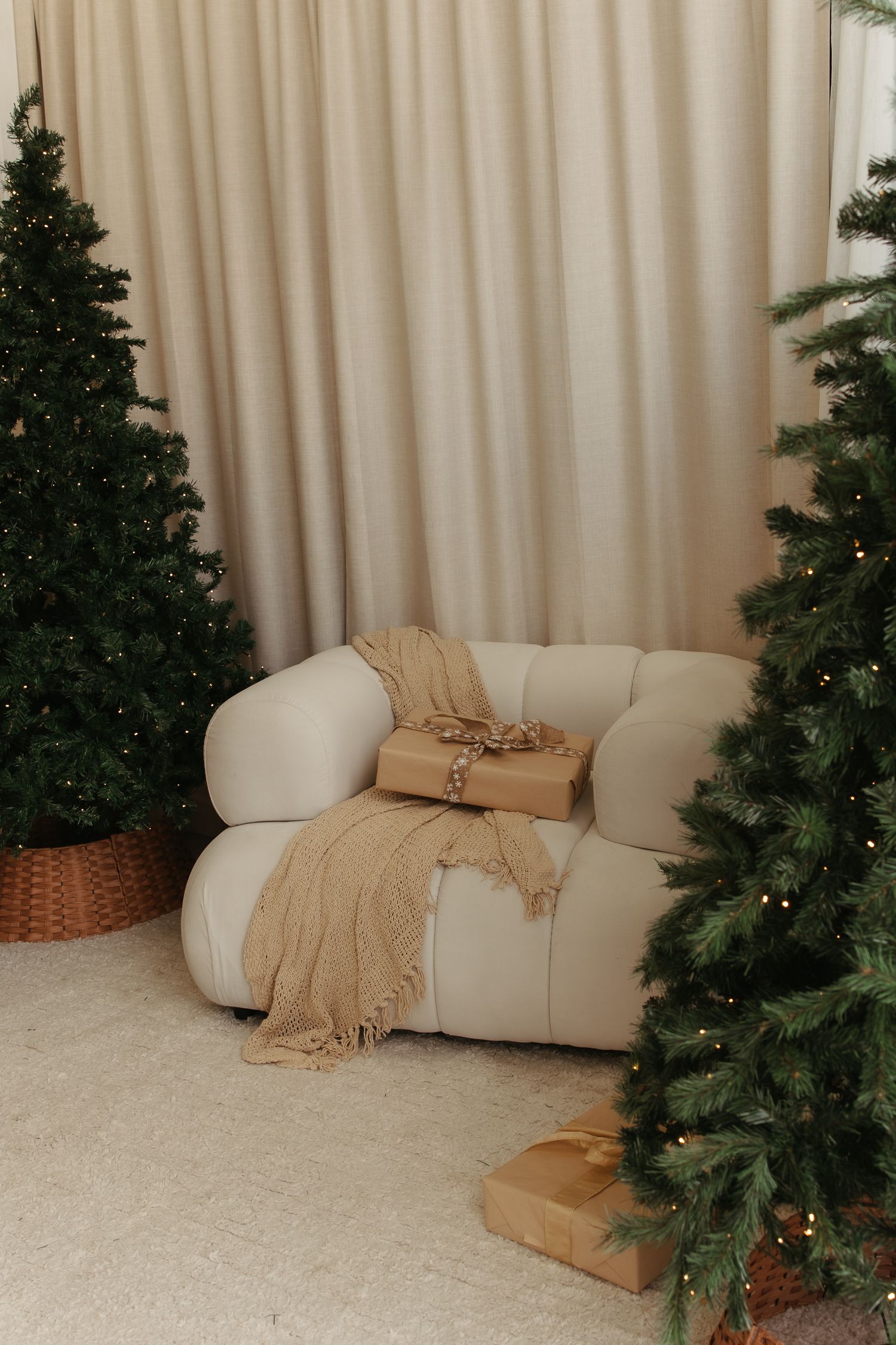 Cozy beige armchair with blanket between decorated Christmas trees and wrapped gifts on white carpet.