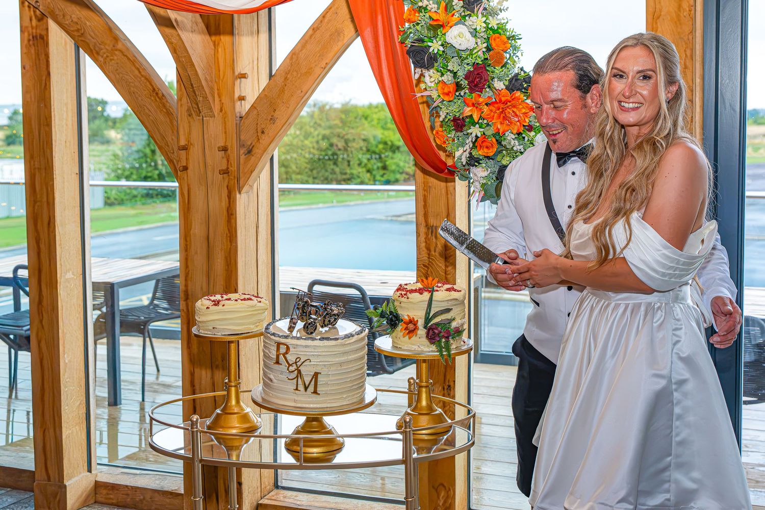 Bride and groom smiling together cutting a monogrammed wedding cake indoors with floral decorations.