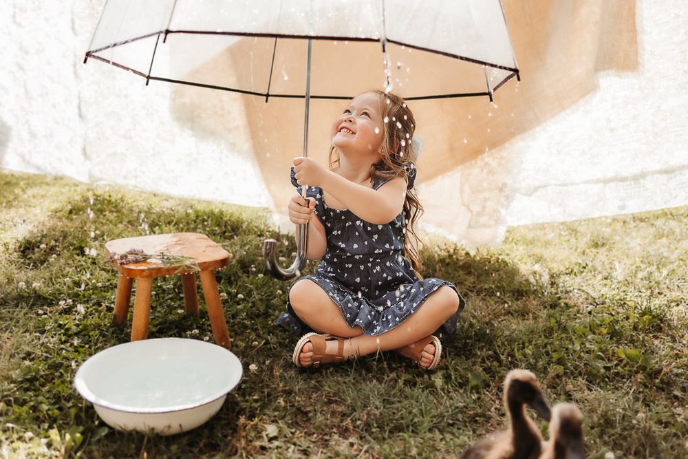 Young girl sitting on grass under clear umbrella with water droplets, beside white basin and wooden stool outdoors.