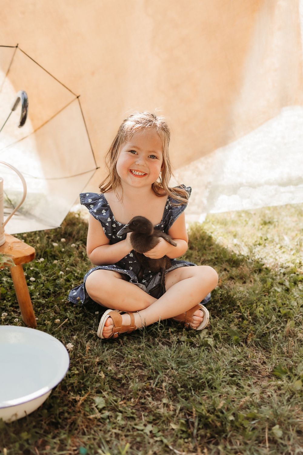 Smiling toddler girl in navy floral romper sitting cross-legged on grass holding a black duckling outdoors.