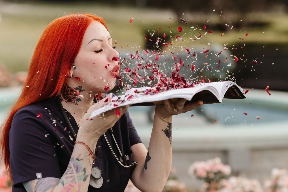 Red-haired tattooed woman blowing red confetti from an open book outdoors near a fountain.