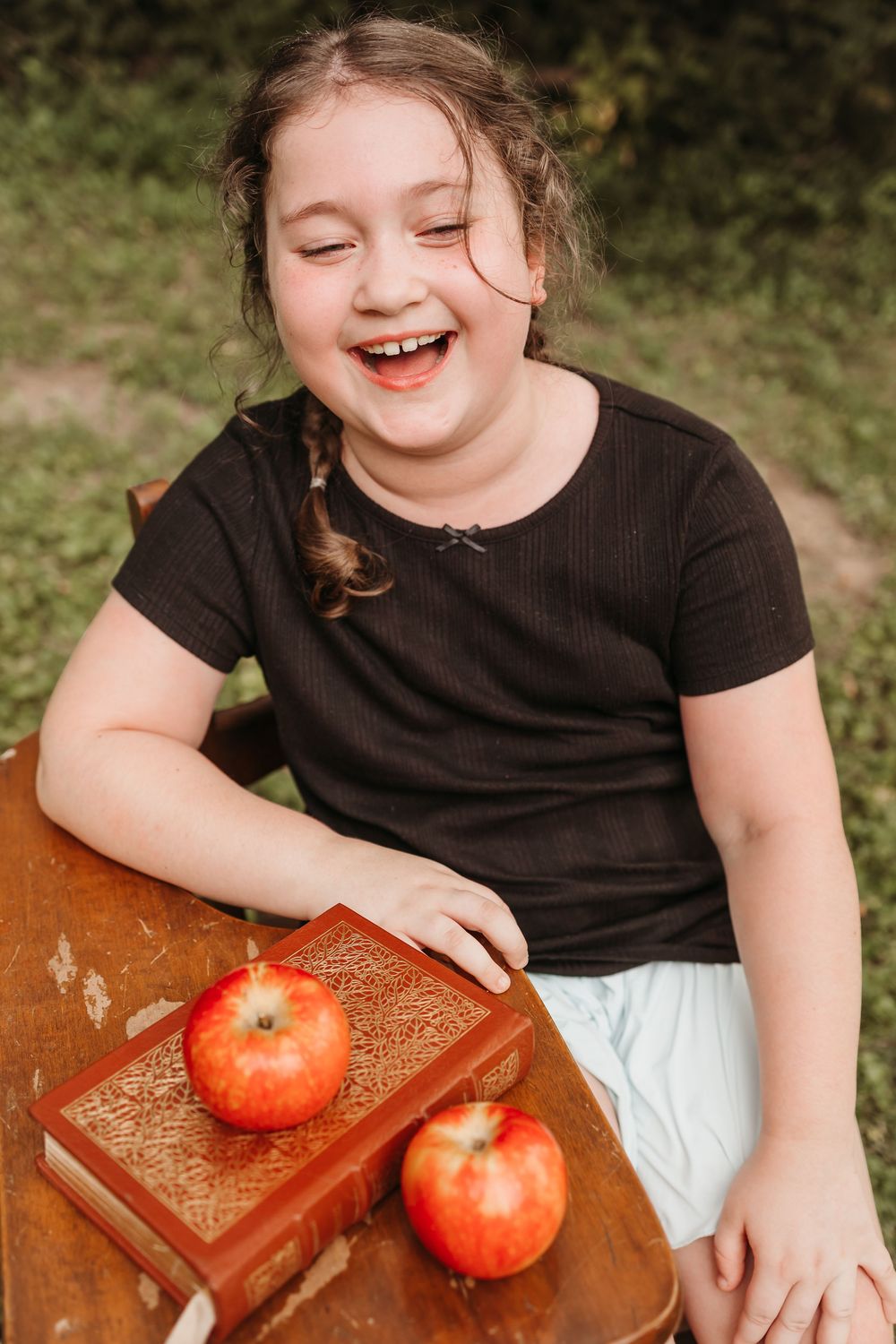 Laughing girl in black shirt sitting outdoors at wooden table with red apples and vintage book.