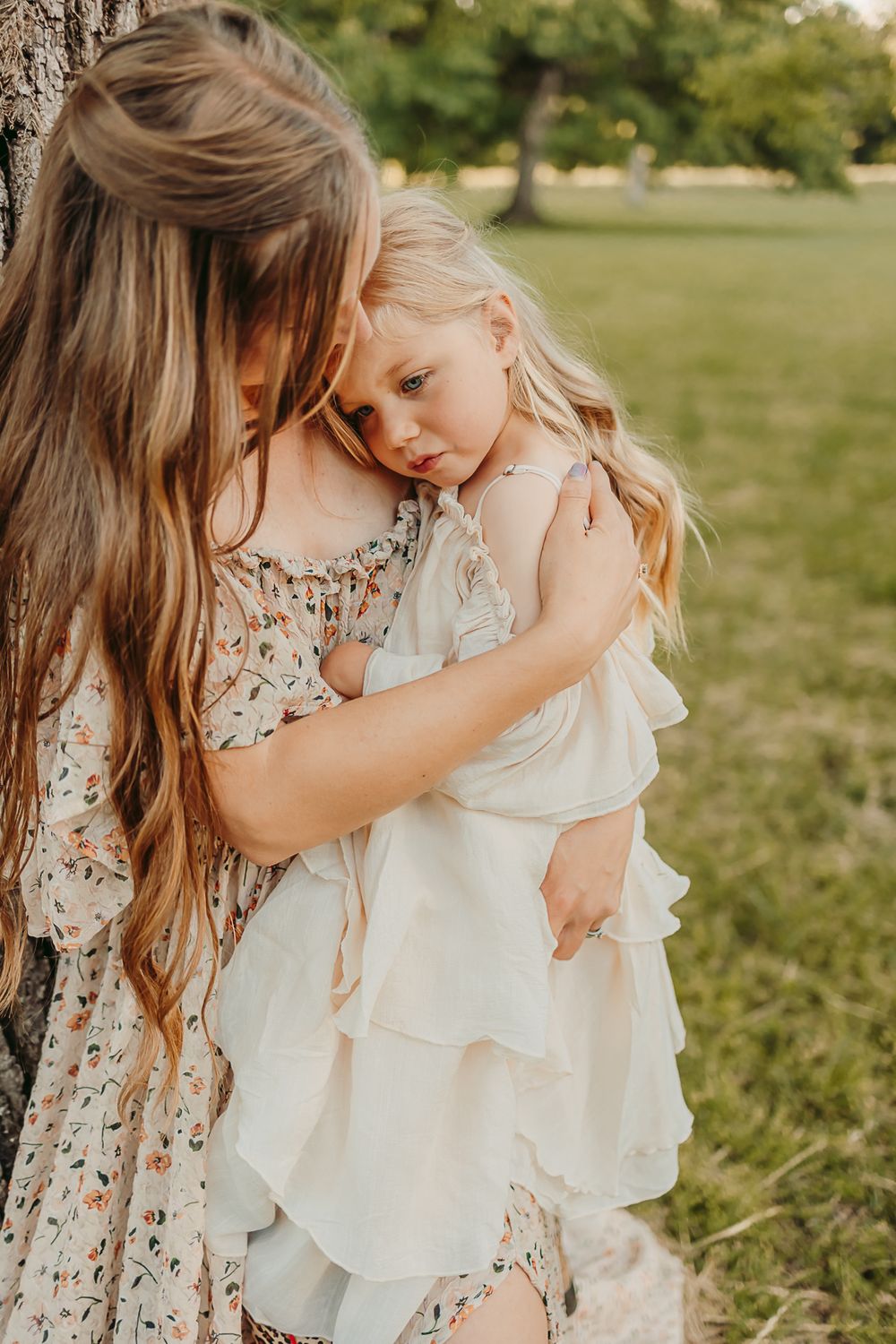 A mother with long brown hair tenderly embraces her young blonde daughter in a white ruffled dress outdoors.