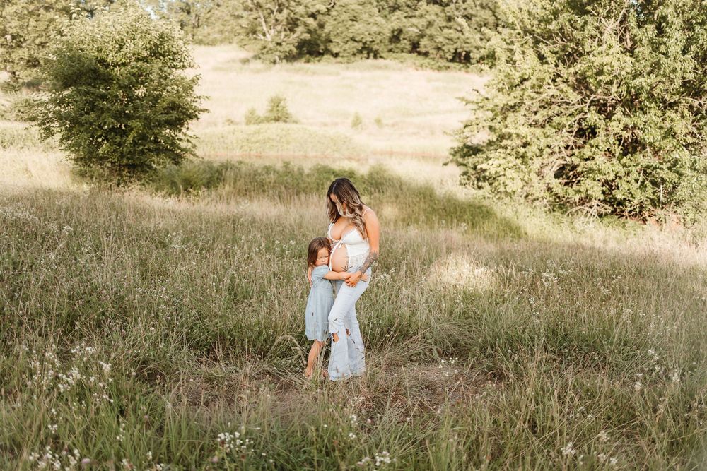 A mother stands in a lush green meadow embracing two young children in soft natural light.