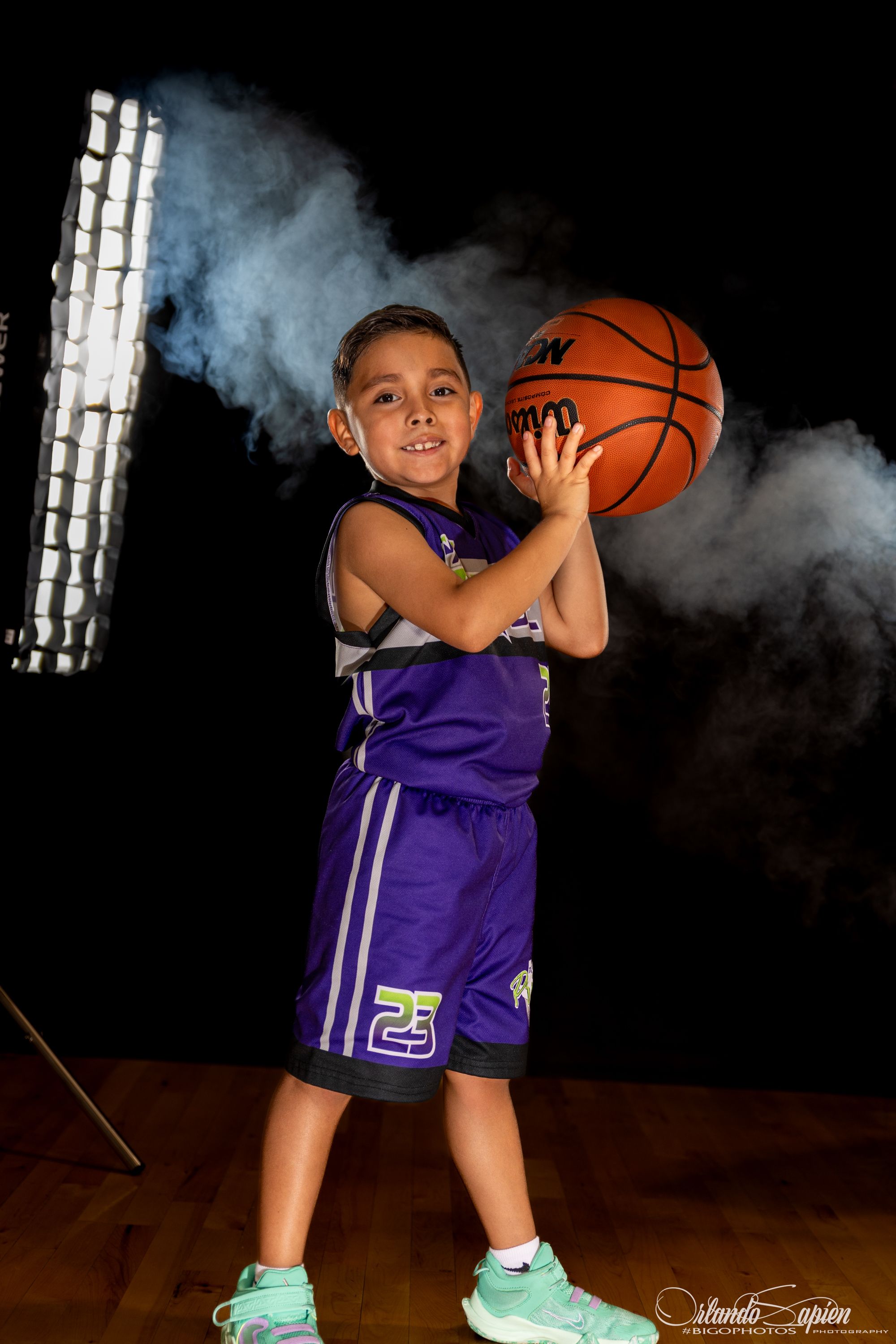 Young boy in purple basketball uniform number 23 holding orange ball posed in dramatic studio lighting with smoke effect.