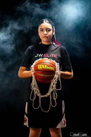 Young female basketball player in JW Elite jersey holds Wilson Evolution ball with net against dark smoky background.
