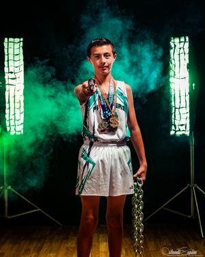 Young athlete in white basketball uniform proudly displaying multiple gold medals and trophies against dramatic green smoke backdrop.