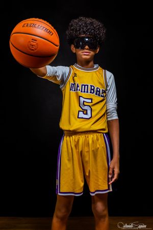Young basketball player in yellow Mambas #5 jersey holding ball, wearing sunglasses against dark background.