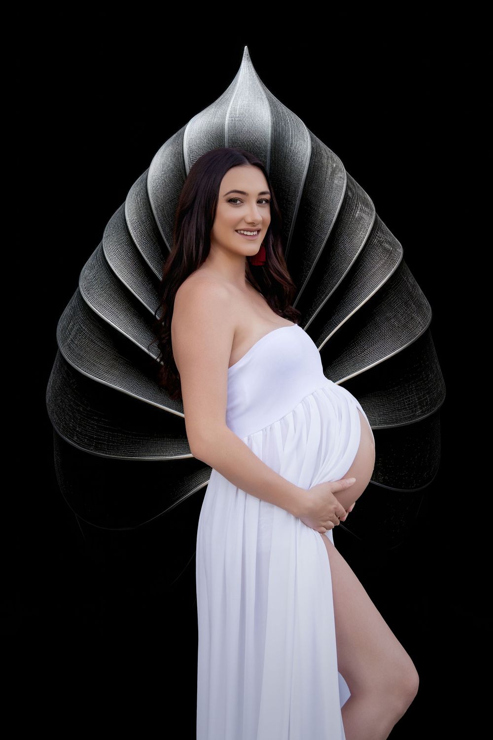 pregnant mother posed in from of a black background with a large shell in the background wearing a white sheer dress