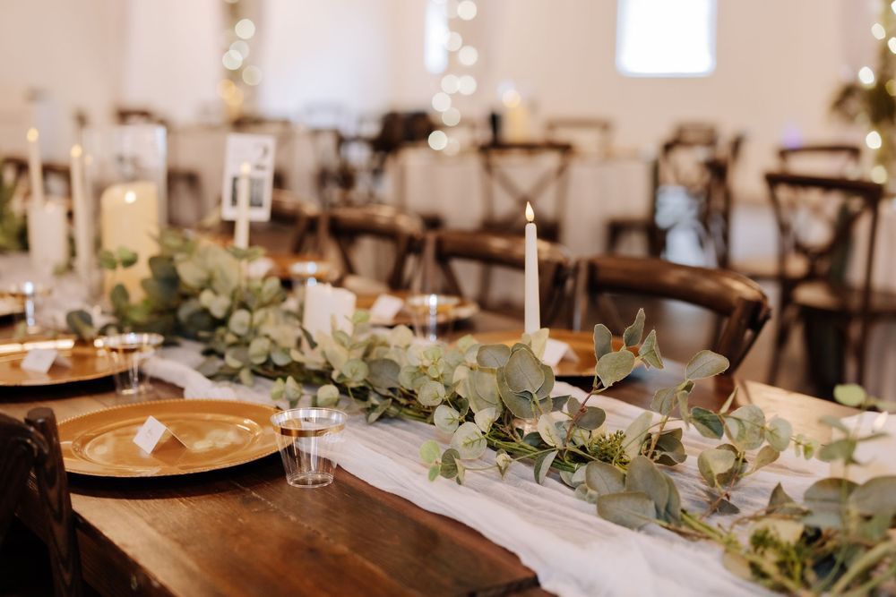 Elegant wedding reception table with eucalyptus garland, white candles, gold chargers, and string lights in a rustic venue.