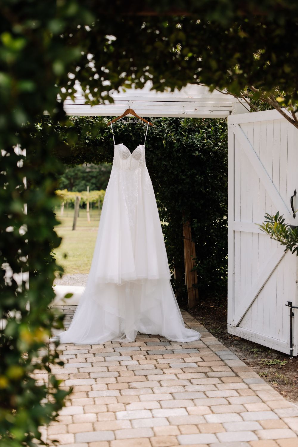 White wedding dress hanging on a wooden hanger in a garden archway with white barn doors.