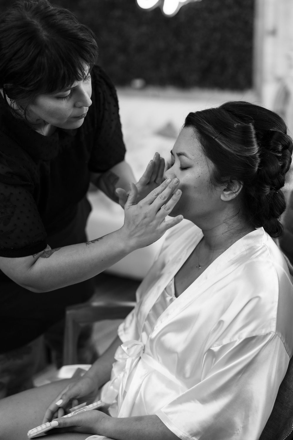 Black and white photo of a makeup artist applying foundation to a bride in a satin robe before her wedding.